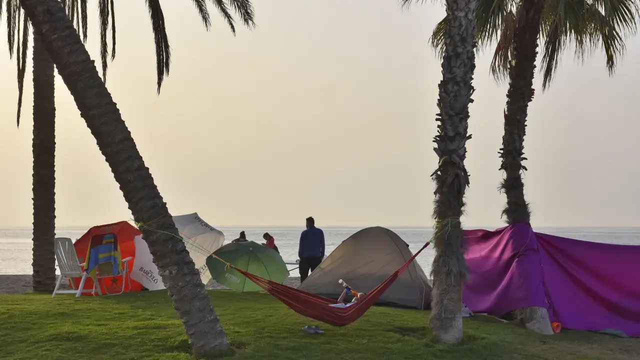Entspannter Camping-Urlaub an der Costa del Sol. Palmen, Zelte, Sonnenschirme und eine Hängematte am Strand.