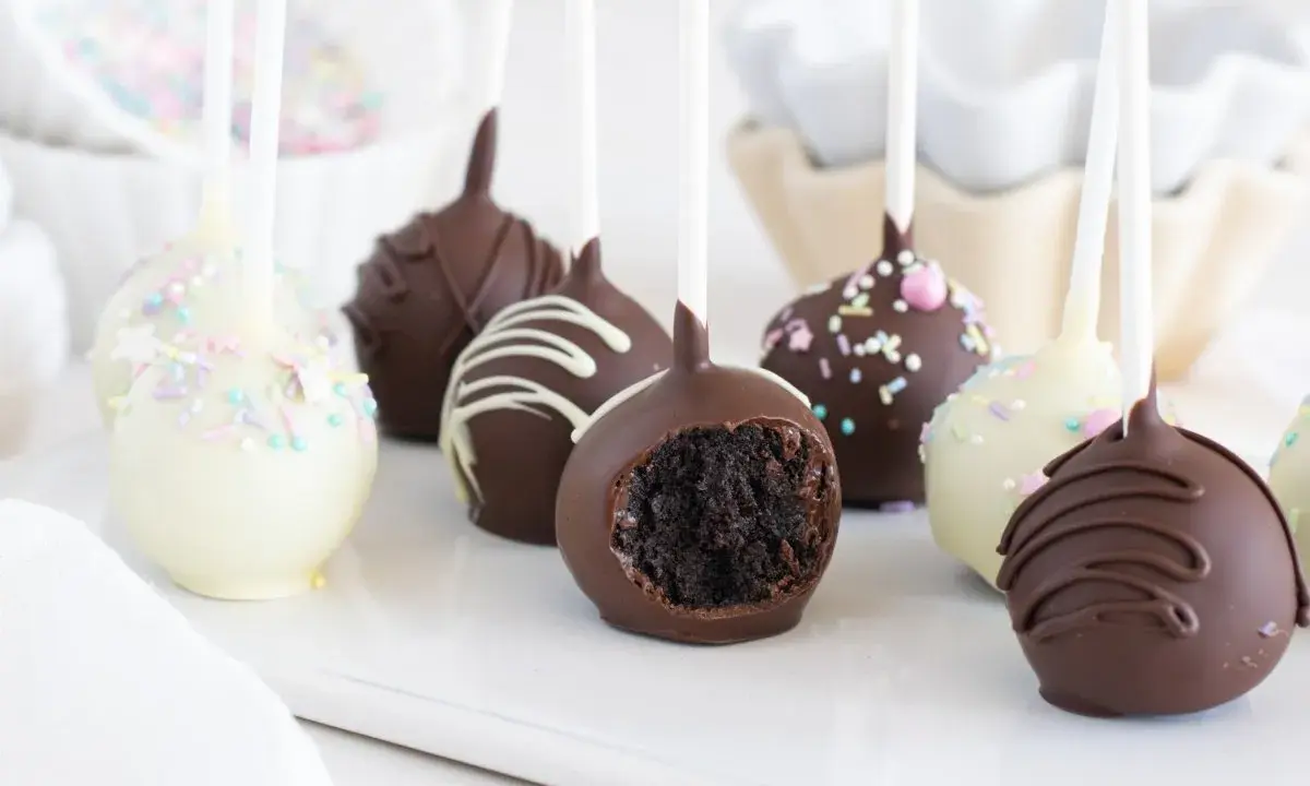 Deliciosos cake pops cubiertos de chocolate, algunos con chispas de colores y otros con glaseado blanco. Uno muestra su interior esponjoso.