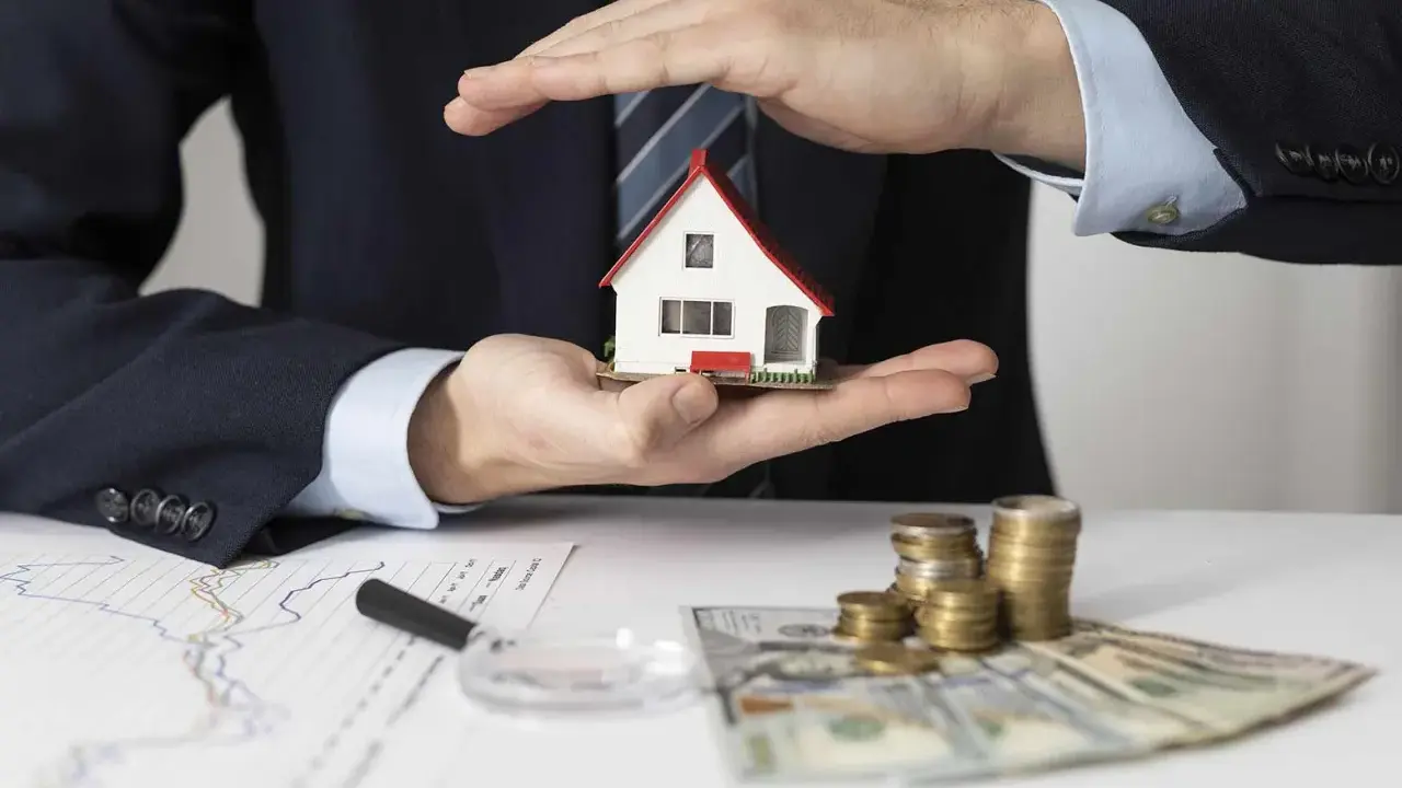 Manos protegen una casa modelo sobre un contrato de alquiler con avalista, junto a dinero y gráficos.