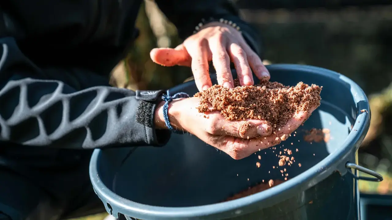 Ręce trzymają zanętę Traper Feeder nad wiaderkiem.