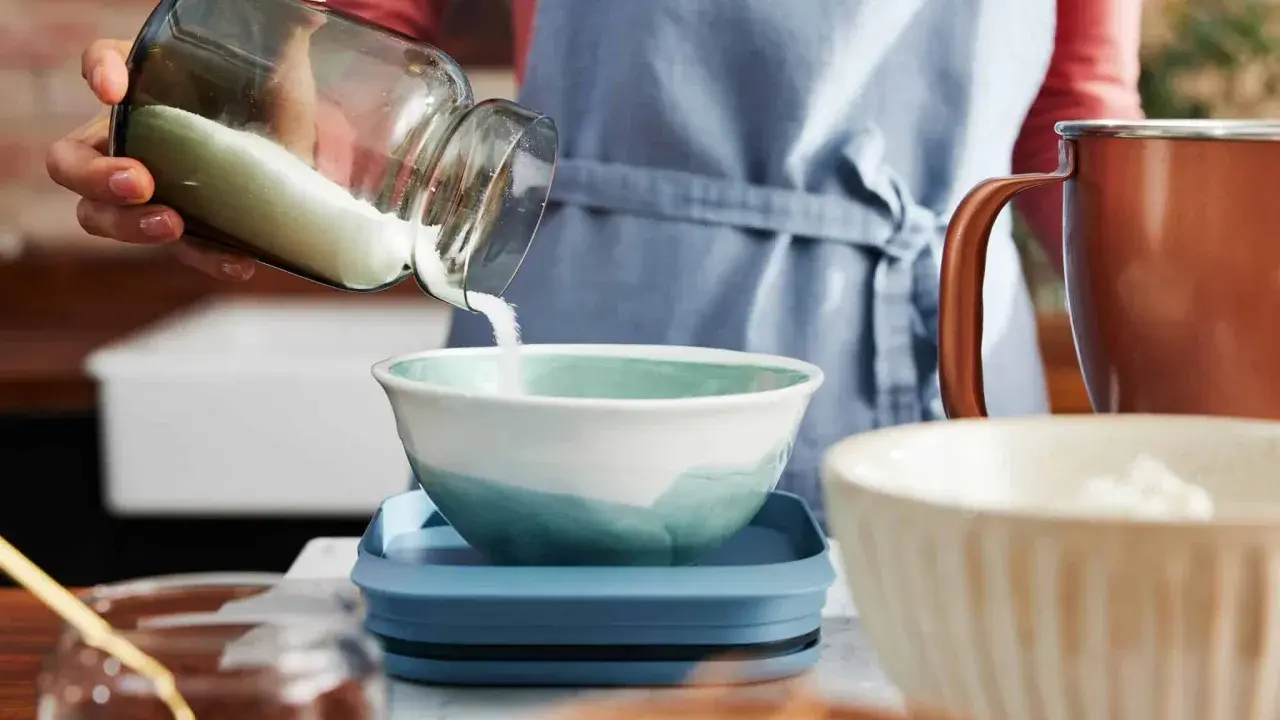 Eine Hand schüttet Mehl aus einem Glas in eine Schüssel. Die Frage, wie viel Gramm Mehl in eine Tasse passen, wird hier visuell thematisiert.