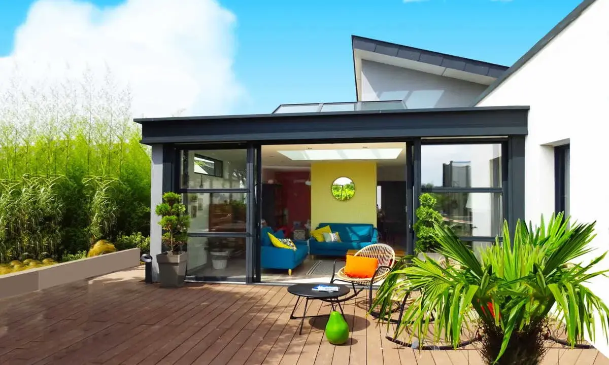 Une véranda moderne avec de grandes baies vitrées s'ouvrant sur une terrasse en bois. Un salon coloré est visible à l'intérieur.