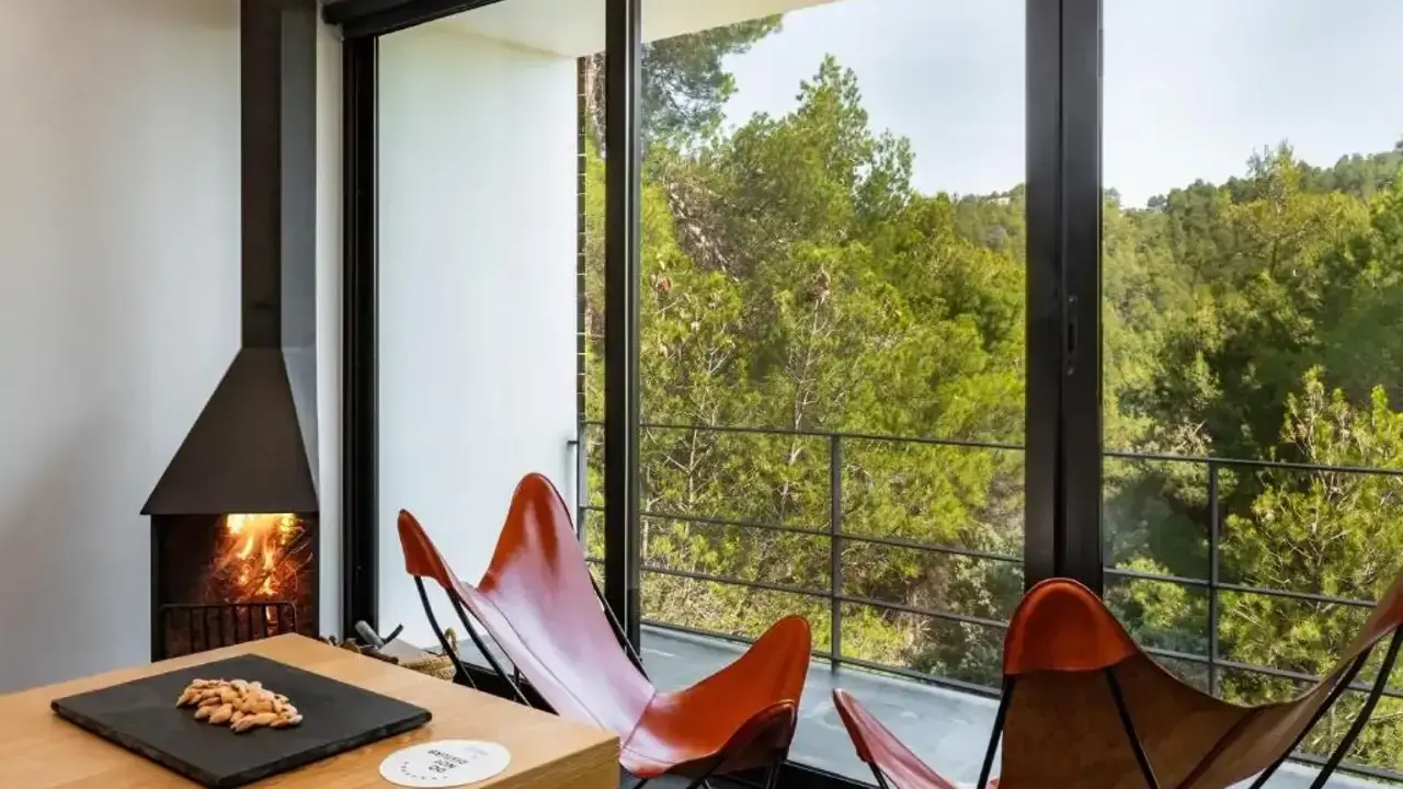 Chimenea encendida y sillones de cuero frente a un balcón con vistas a un bosque frondoso, ideal para el turismo interior España.