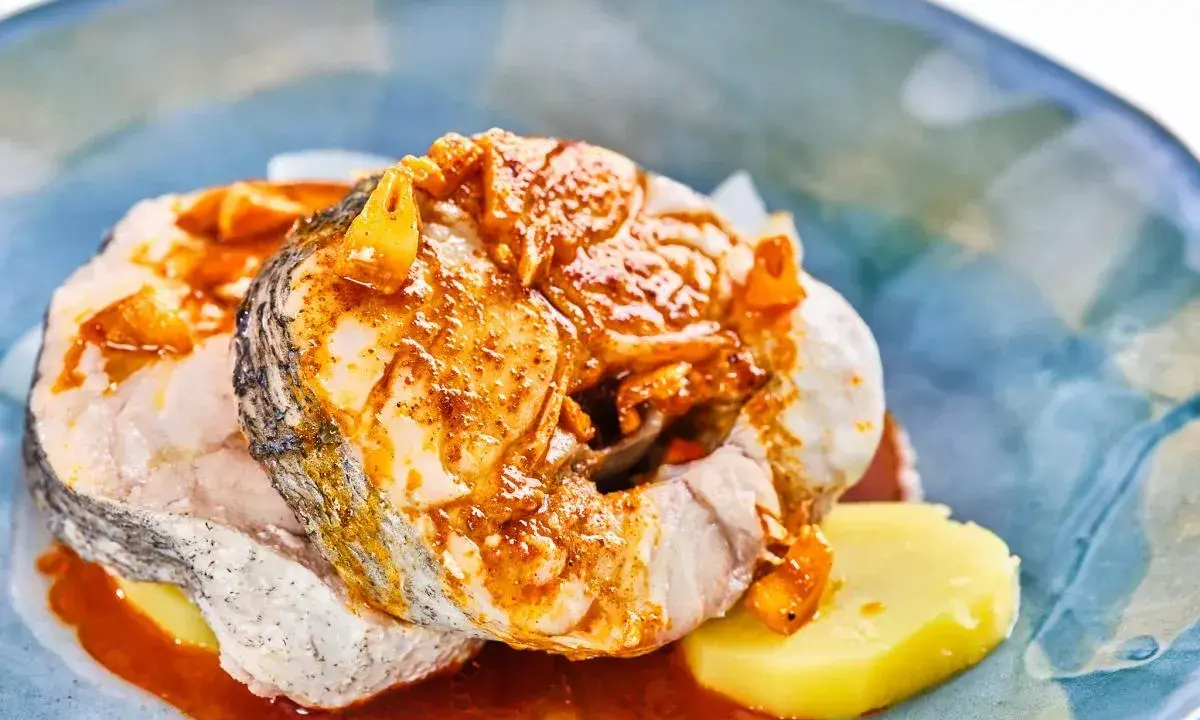 Merluza a la gallega con patatas y salsa de pimentón, servida en un plato azul.