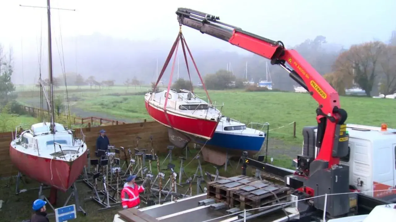 Grue soulevant un bateau rouge pour son recyclage. Un autre bateau bleu est posé sur des supports. L'opération de recyclage bateau se déroule dans un paysage brumeux.