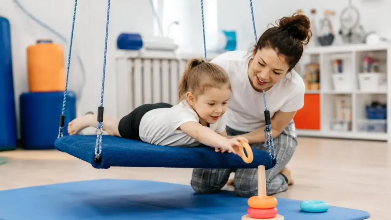Terapia integracji sensorycznej: dziewczynka na huśtawce z terapeutką, która pomaga jej sięgnąć po zabawkę.