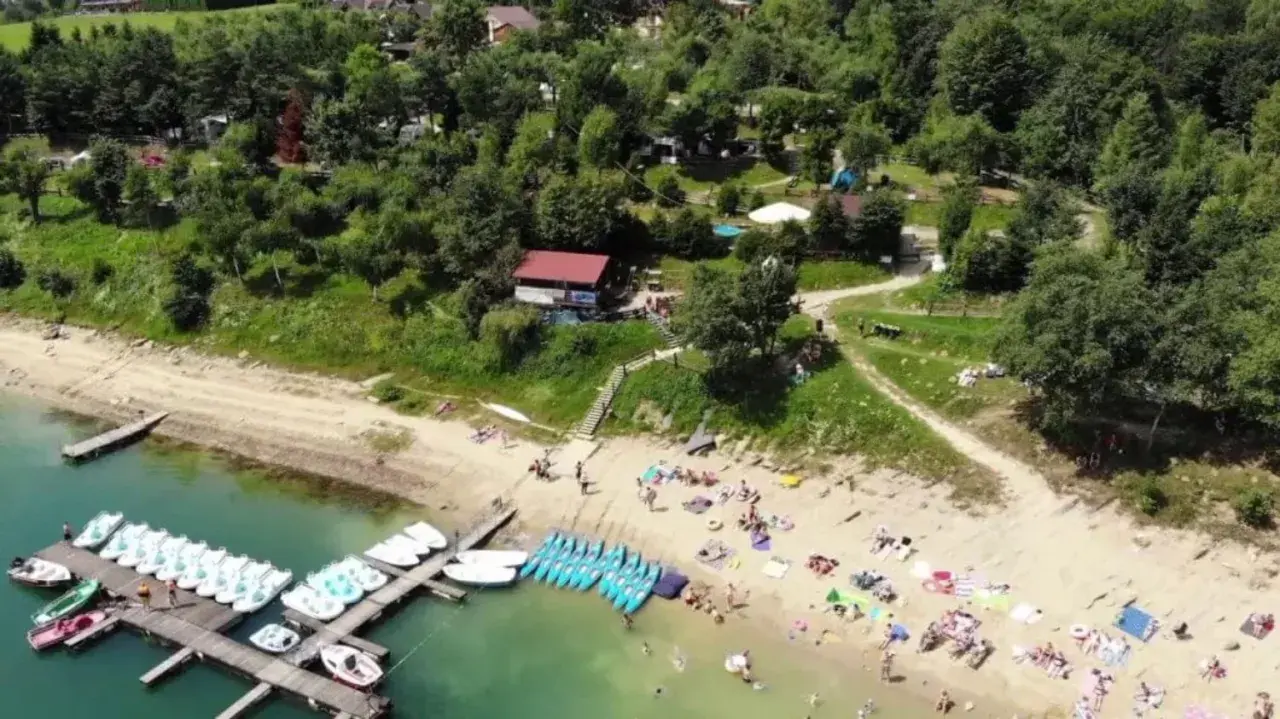 Słoneczna plaża w Polańczyku tętni życiem. Ludzie odpoczywają na piasku, a w wodzie pływają rowery wodne i kajaki.