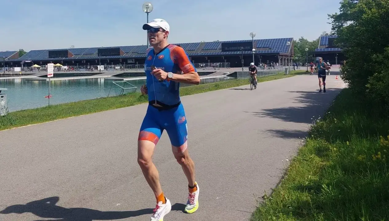Triathlet im blauen Anzug läuft auf einer Straße. Er fragt sich, wie wird man schneller. Ein Radfahrer ist im Hintergrund zu sehen.