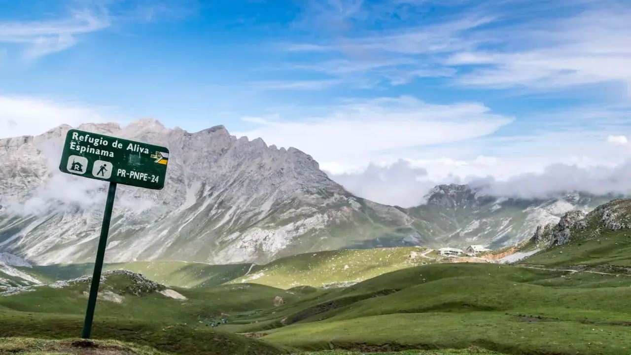 Wanderwegschild weist den Weg zum Refugio de Aliva und Espinama in den **Lagos de Covadonga**. Grüne Hügel und zerklüftete Berge unter blauem Himmel.