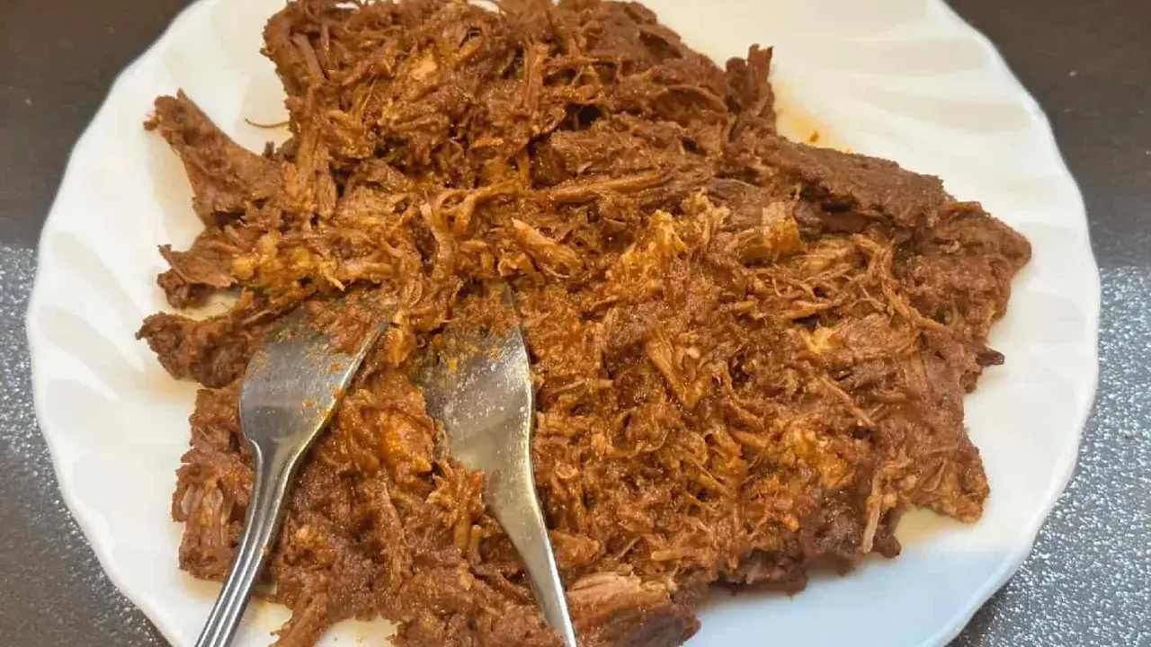 Delicioso pulled pork deshebrado en un plato blanco, listo para ser servido. Dos tenedores se clavan en la carne jugosa.