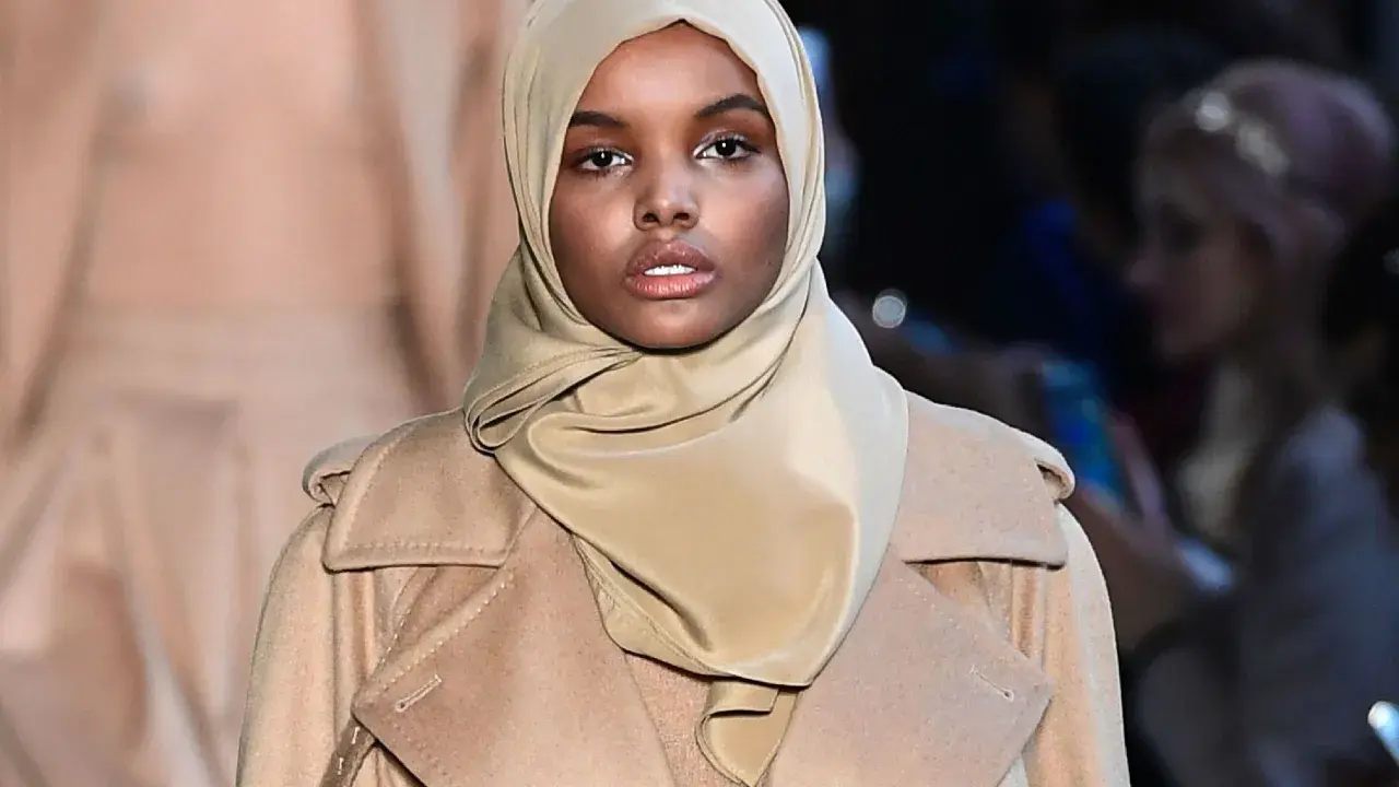 Halima Aden walks the runway in a stylish beige coat and hijab, exuding confidence and grace.