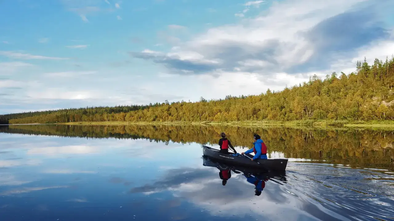 Två personer paddlar kanot på en spegelblank sjö i Sverige. Skogen längs stranden reflekteras i vattnet.