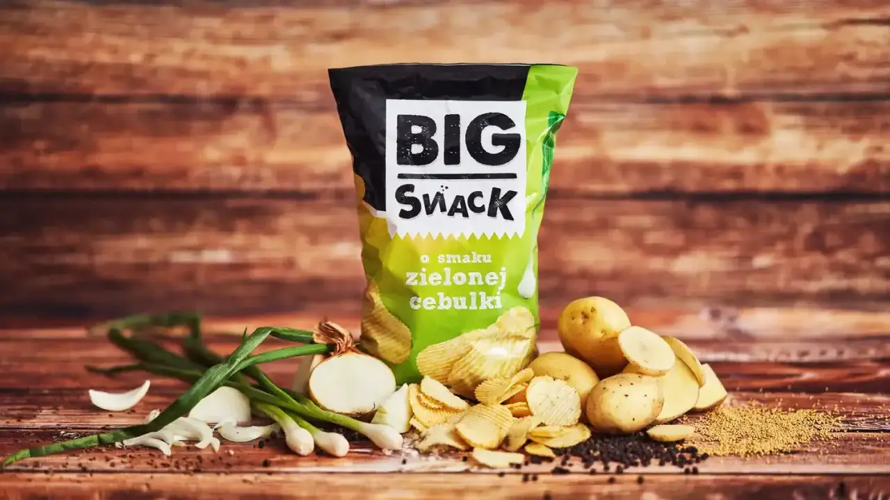 Reklamowa fotografia paczki chipsów "Big Snack" o smaku zielonej cebulki, z ziemniakami i cebulką w tle.