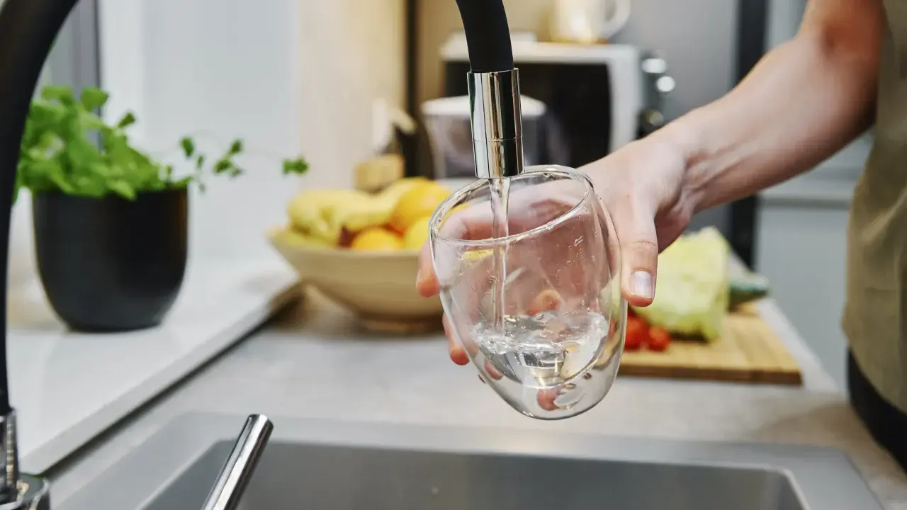 Ręka nalewa wodę do szklanki. W tle widać owoce i warzywa, jakby przygotowywano posiłek. Czysta woda, jak w akwarium.