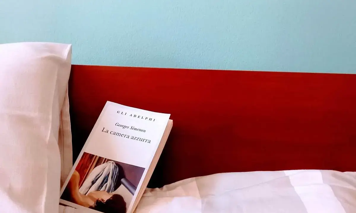 Un libro aperto, "La camera azzurra" di Simenon, giace su un letto bianco, pronto per essere letto.