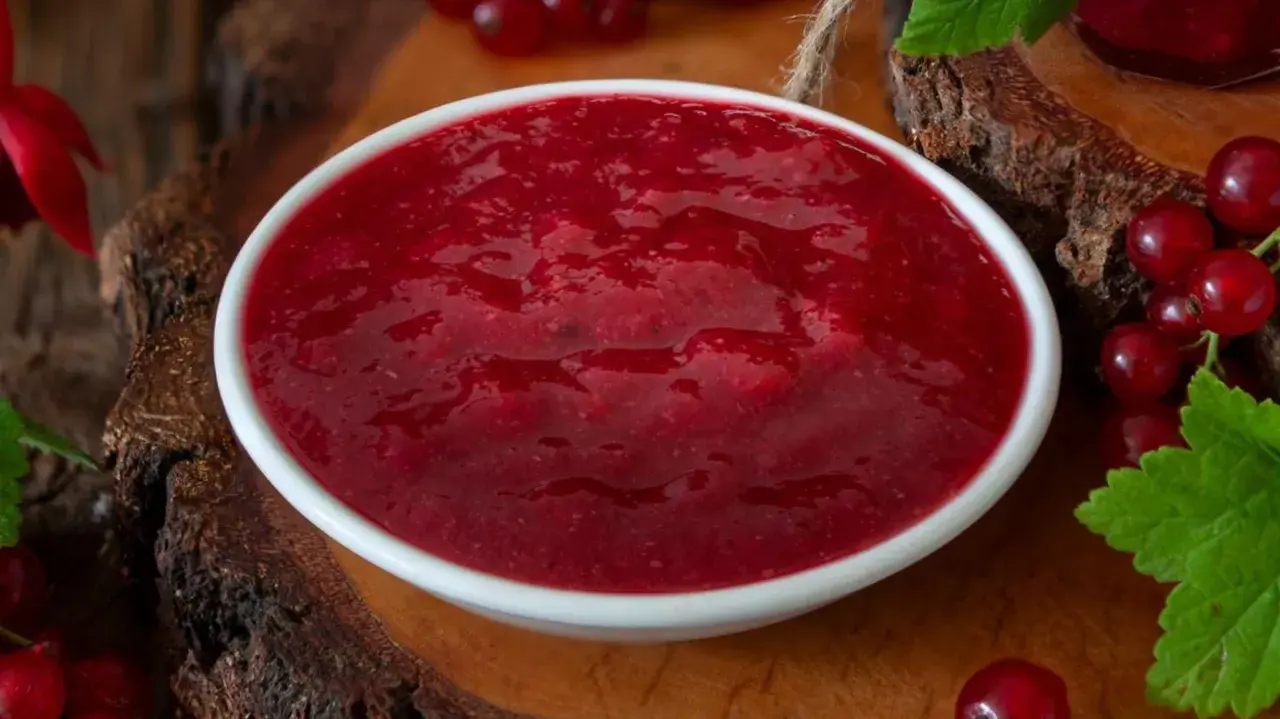 Eine Schüssel mit leuchtend roter Cumberland Sauce, umgeben von frischen Johannisbeeren und grünen Blättern.