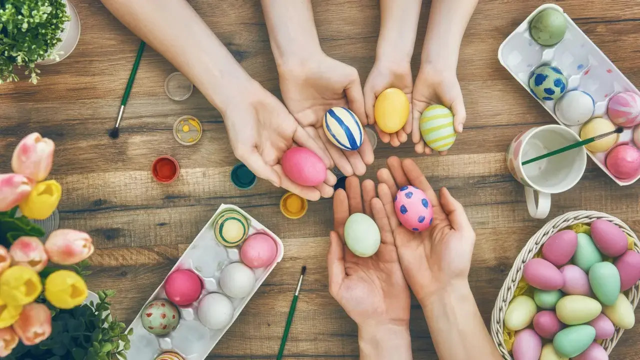 Hände von Kindern und Erwachsenen bemalen bunte Ostereier. Ein Korb voller Eier und Tulpen auf Holztisch.