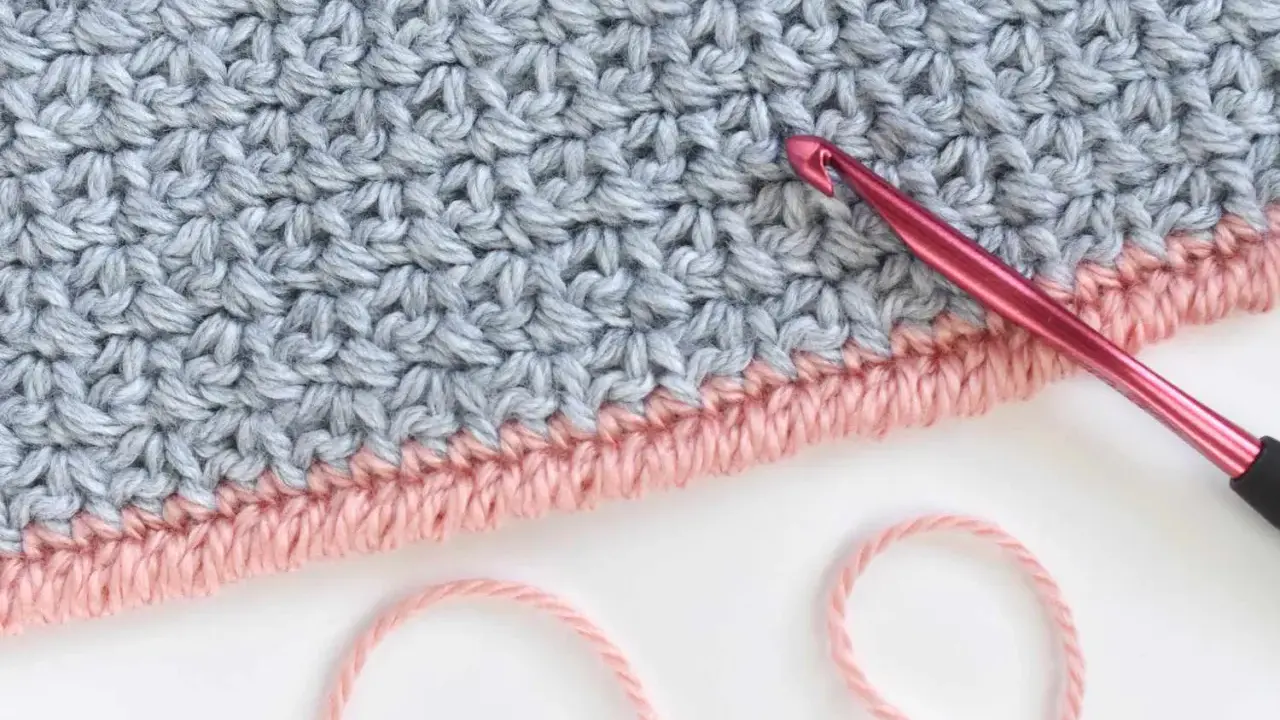 Detalle de tejido a crochet con punto cangrejo en rosa y gris. Una aguja rosa está lista para continuar el trabajo.