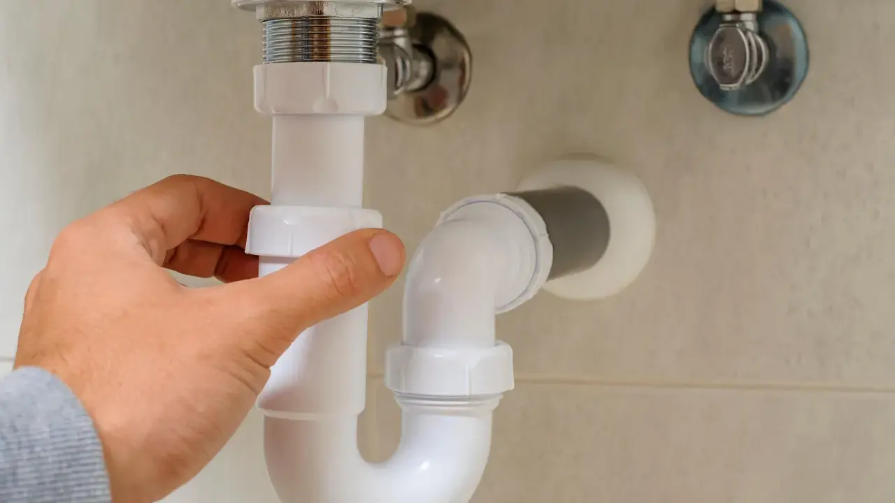 Mano ajustando la altura del desague del lavabo, tubería blanca de PVC bajo el lavamanos.