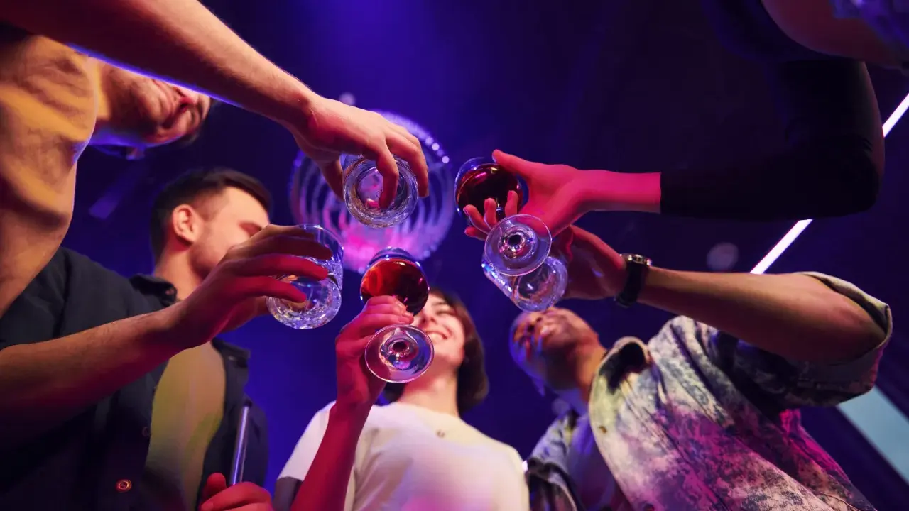 Grupa przyjaciół wznosi toast, świętując weekend. W tle dyskotekowe światła, sugerujące zabawę i być może początek weekendowego alkoholizmu.