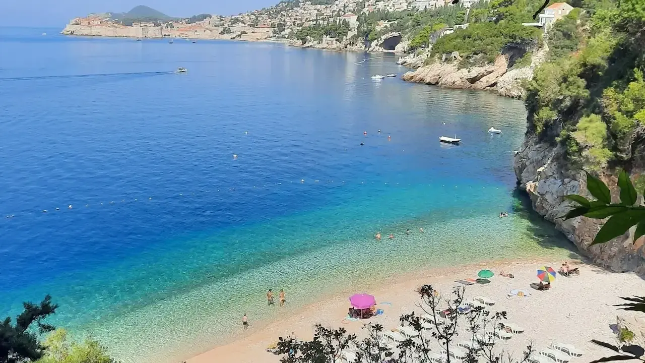 Malownicza zatoka z turkusową wodą i kamienistą plażą. W oddali widać mury Starego Miasta Dubrownik. Idealne plaże Dubrownik na relaks.