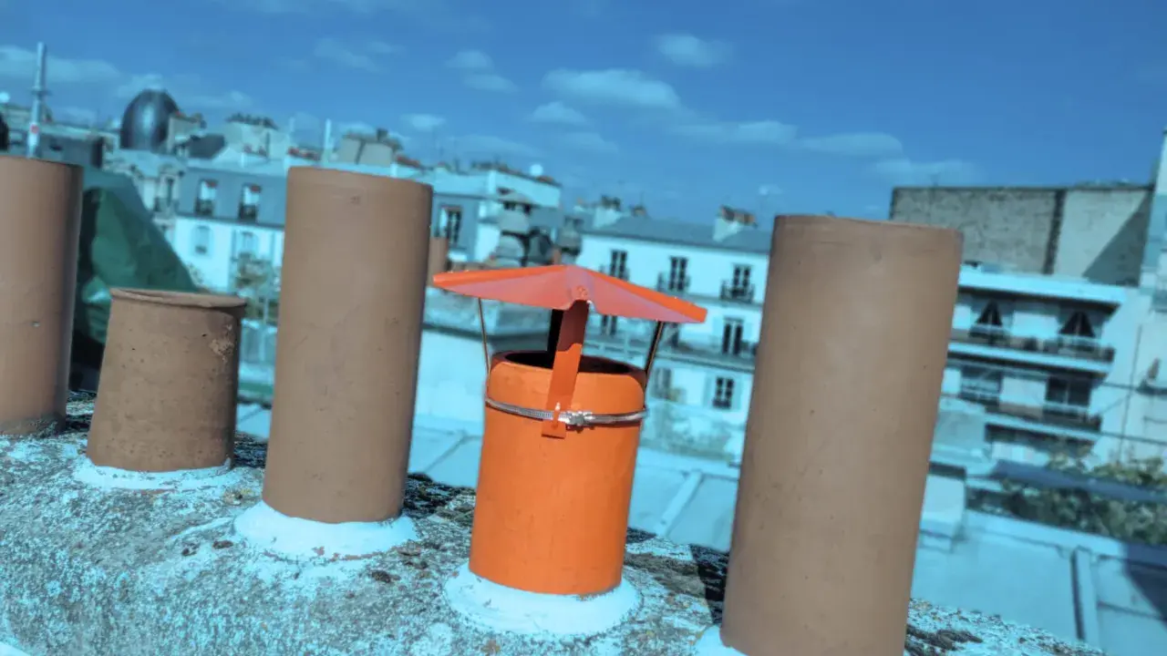 Sur un toit parisien, un chapeau chinois orange protège une cheminée, entouré d'autres conduits sous un ciel bleu.