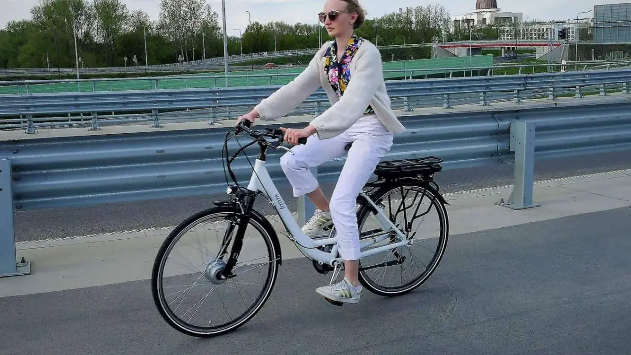 Młoda kobieta w białych spodniach i jasnym swetrze jedzie na rowerze elektrycznym, ciesząc się jazdą i osiągając swoją maksymalną prędkość.