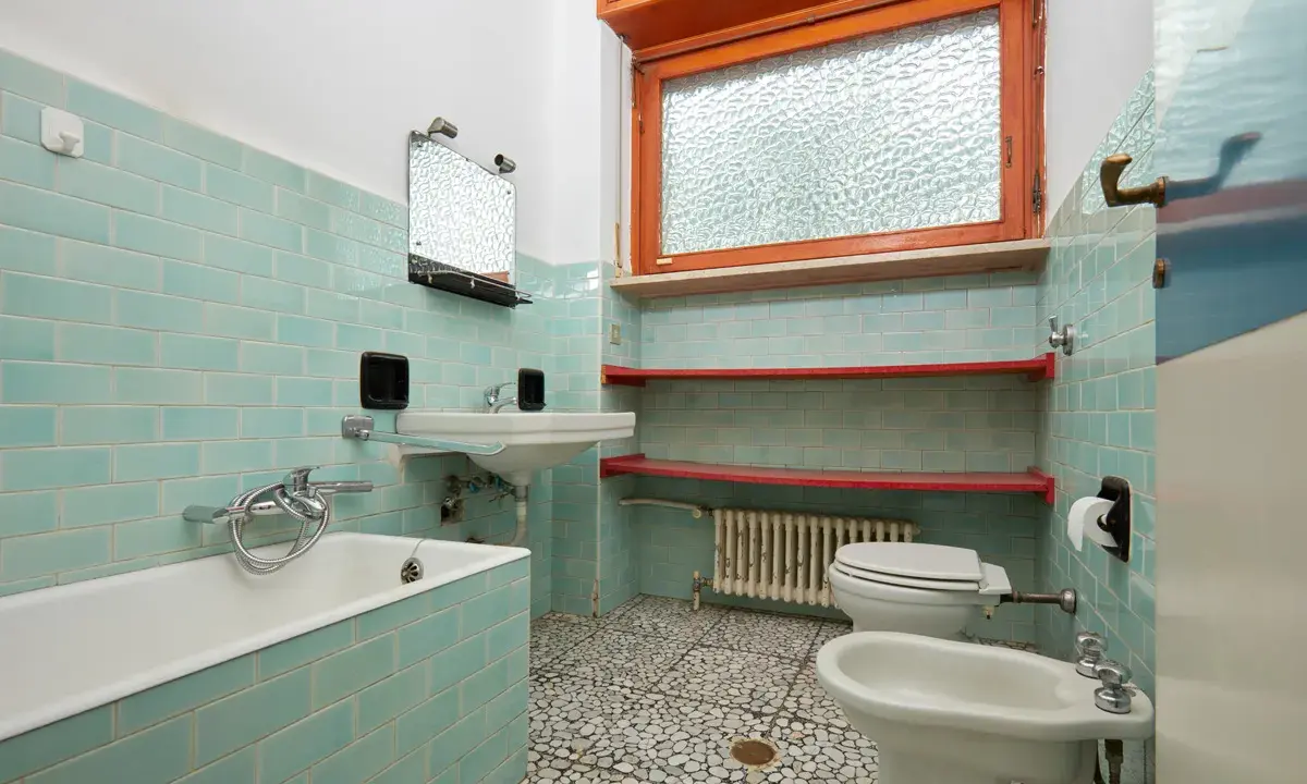 Bad renovieren ohne fliesen abschlagen: hellblau geflieste Wände, Badewanne, Waschbecken, Toilette, Bidet und Fenster.