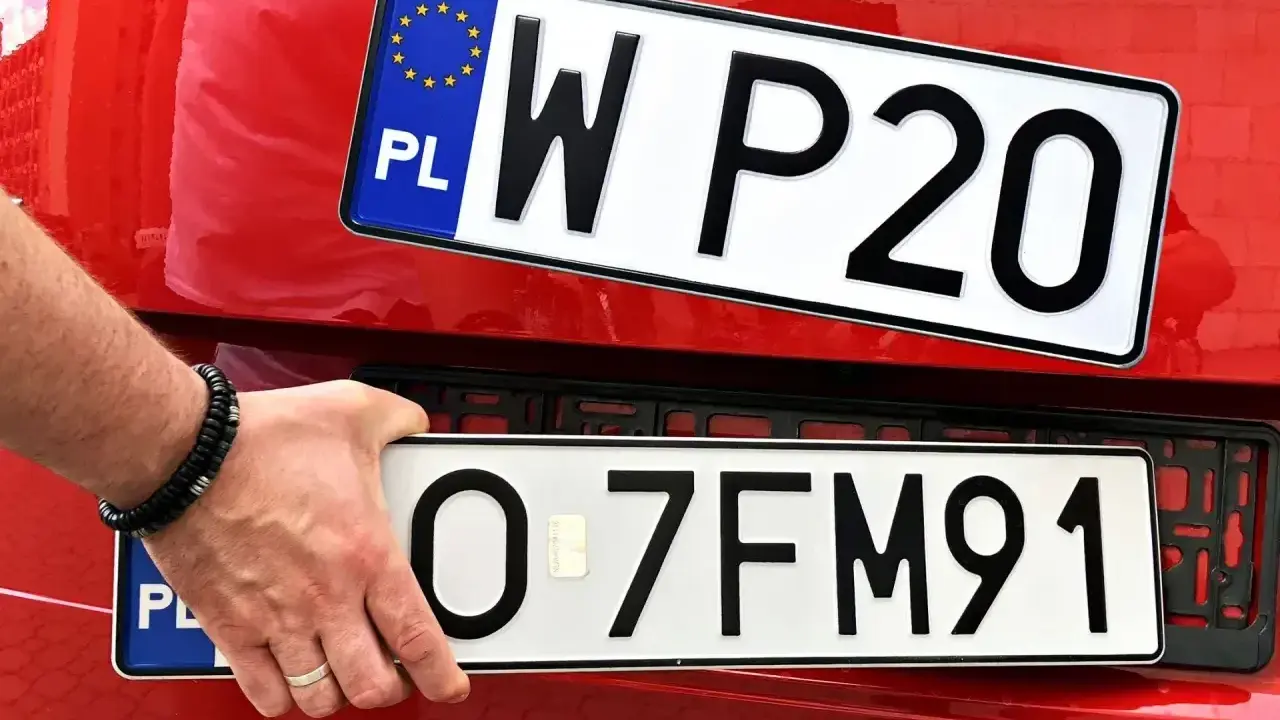 Dłoń z bransoletką montuje tablice rejestracyjne na czerwonym samochodzie. Widać dwie tablice: jedną z napisem WP20, drugą z numerem O-7FM91.