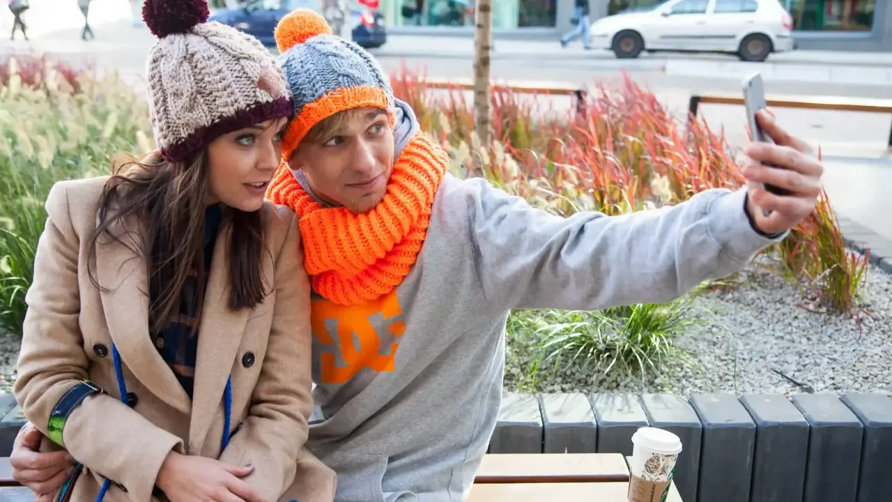 Para do selfie w zimowych stylizacjach. Ona w beżowym płaszczu i czapce z pomponem, on w szarej bluzie i czapce z pomarańczowym akcentem. Jaka czapka do płaszcza? Ta jej wygląda świetnie!