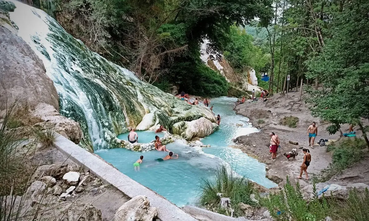 Cascata bianca e pozze azzurre alle terme San Filippo, con persone che si rilassano nell'acqua termale.