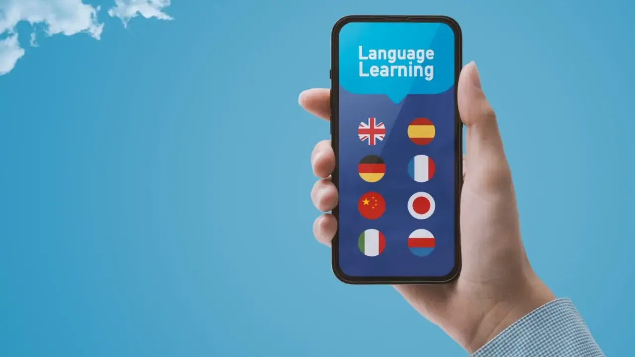Dłoń trzyma smartfon z aplikacją do nauki języków. Wybór między Busuu czy Duolingo? Na ekranie flagi: UK, Hiszpanii, Niemiec, Francji, Chin, Japonii, Włoch, Polski.