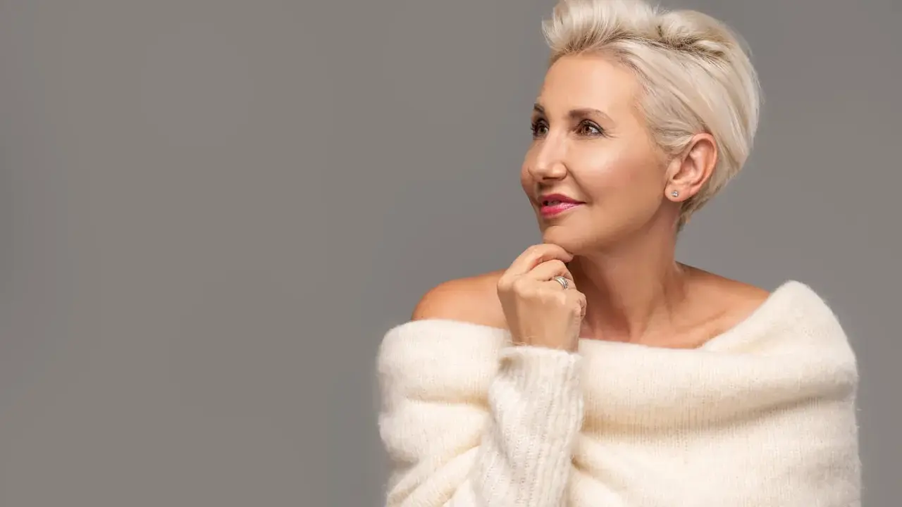 Le visage d'une femme de 80 ans, aux cheveux blonds courts, porte un pull blanc en maille. Elle regarde sur le côté, la main sous le menton.