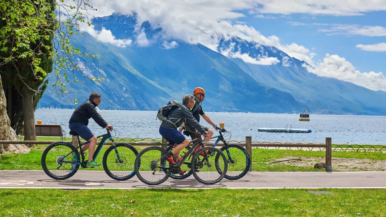 Ciclisti godono di una giornata soleggiata lungo uno dei laghi nord Italia, con montagne maestose sullo sfondo.