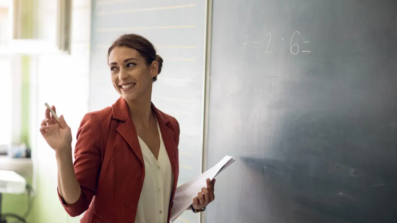 Lehrerin mit Kreide und Unterlagen vor einer Tafel mit einer Matheaufgabe. Sie lächelt und wirkt engagiert, was auf einen interaktiven Unterricht hindeutet.