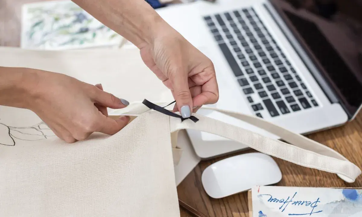 Ręce tworzą torbę DIY, przyszywając czarną wstążkę do płóciennego materiału. Obok leży laptop i myszka.