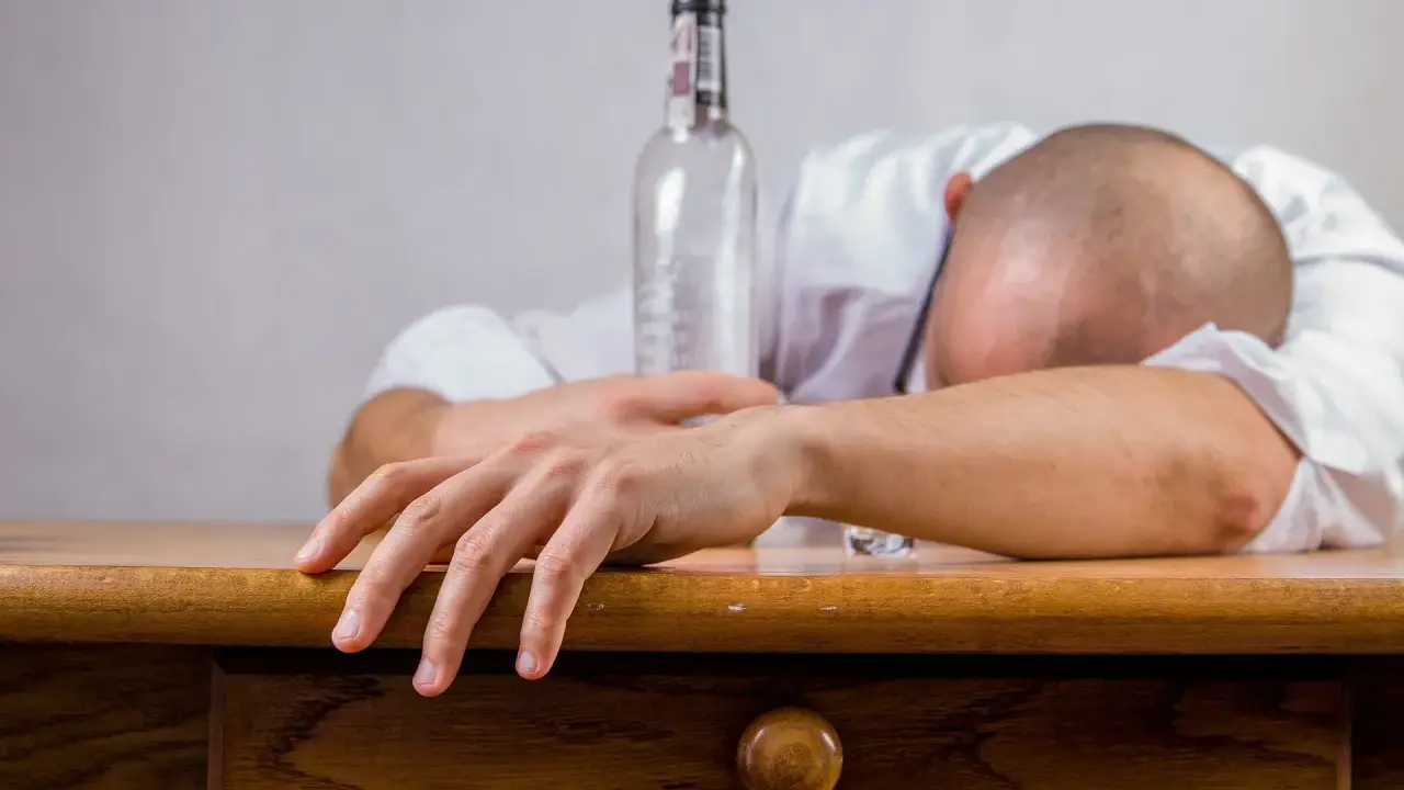 Mężczyzna w białej koszuli leży na stole, głowa oparta na ramieniu. Obok pusta butelka, symbolizująca skutki spożycia alkoholu, jakimi mogą być wymioty po alkoholu.