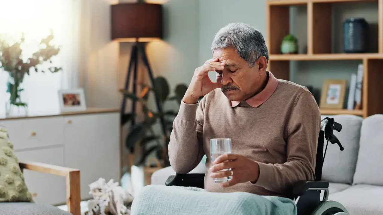 Älterer Mann im Rollstuhl mit Kopfschmerzen, hält ein Glas Wasser. Möglicherweise ein Parkinson-Test der Haut, der auf seine Symptome hinweist.
