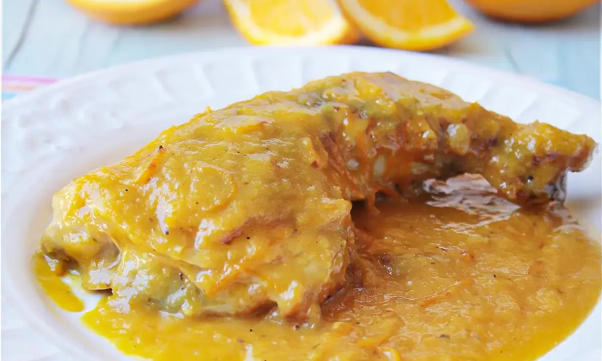 Delicioso pollo a la naranja, bañado en una salsa dorada y jugosa, con gajos de naranja frescos al fondo.