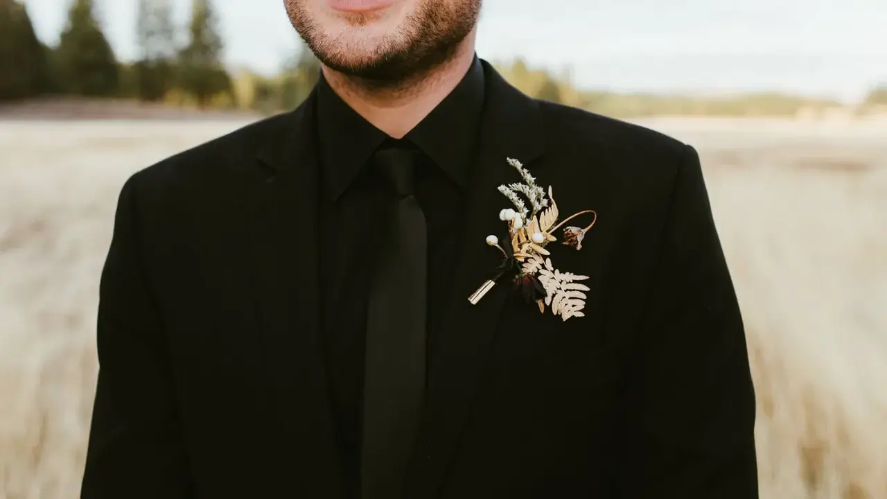 A groom in all black wedding guest attire men's suit, complete with a black tie and a rustic boutonniere.