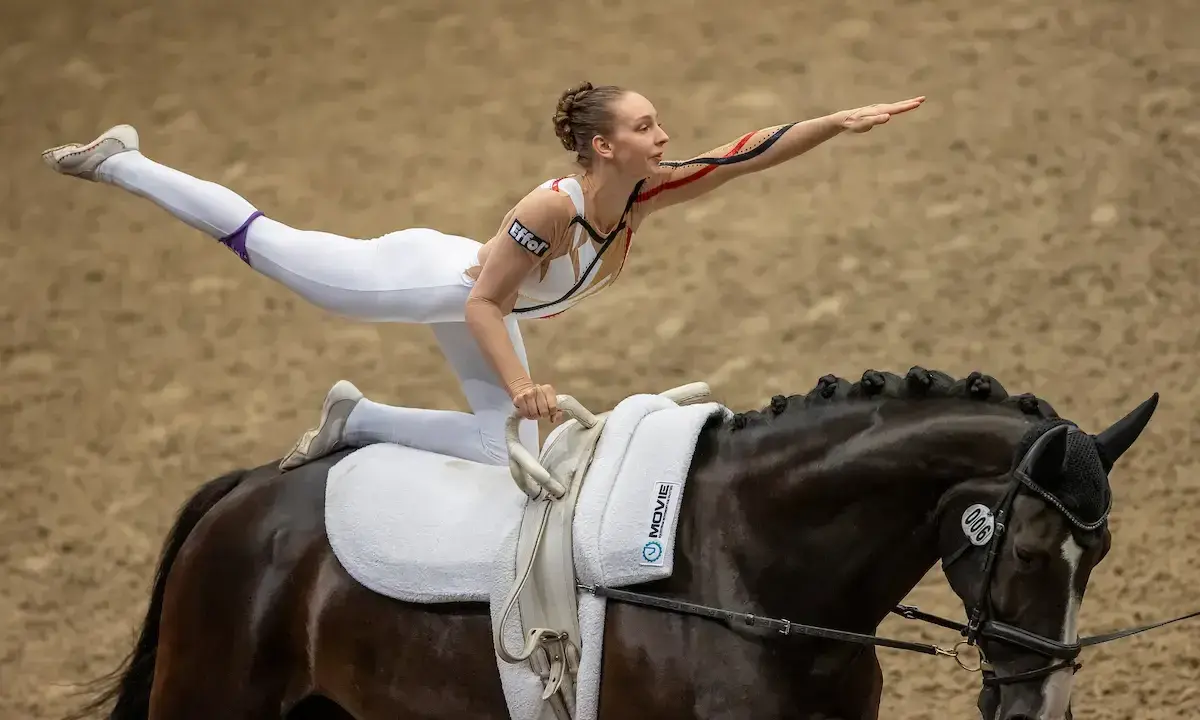 Eine Voltigiererin in weißem Anzug balanciert auf einem dunklen Pferd, bereit für das Voltigiieren Olympia.