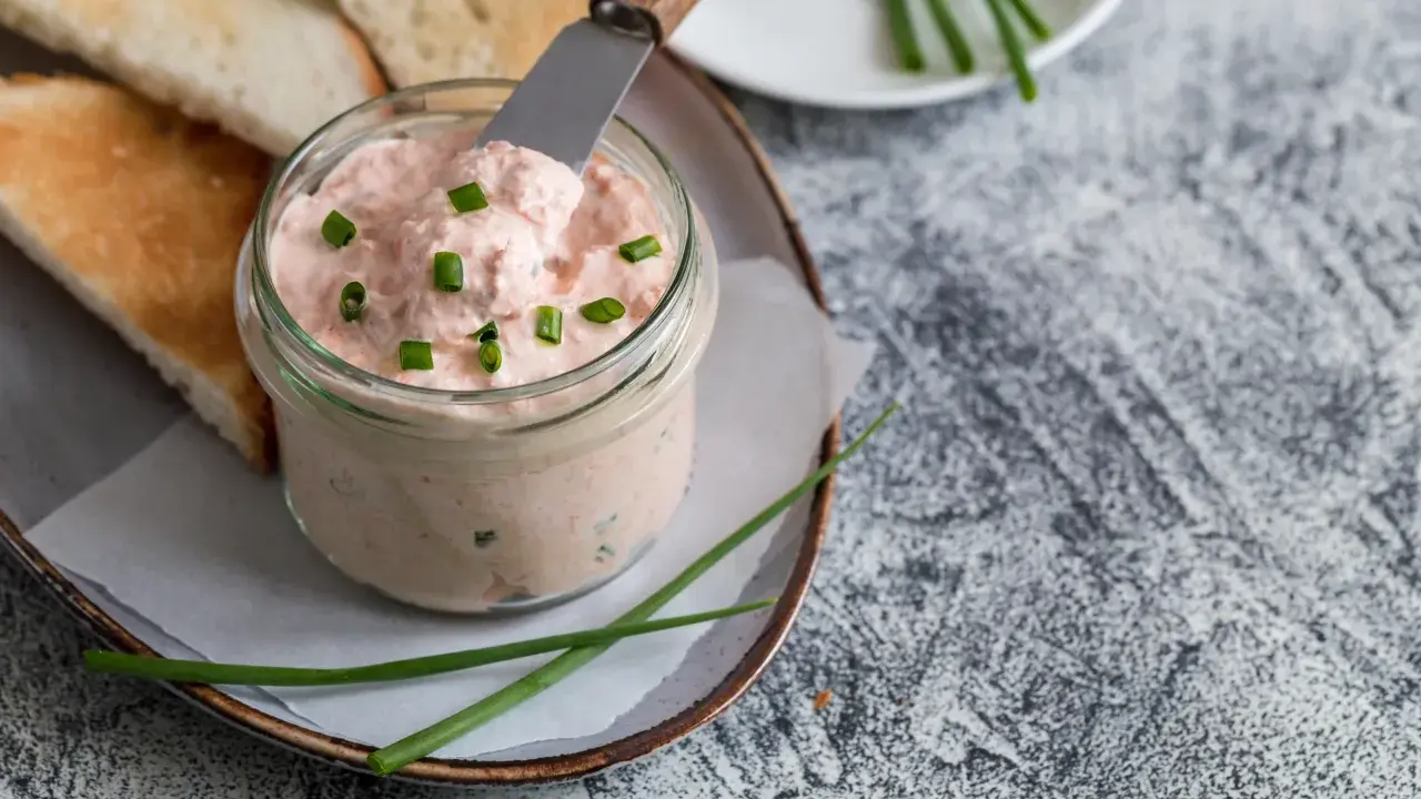 Un pot de rillettes de saumon crémeuses, garni de ciboule, prêt à être tartiné sur des toasts grillés.
