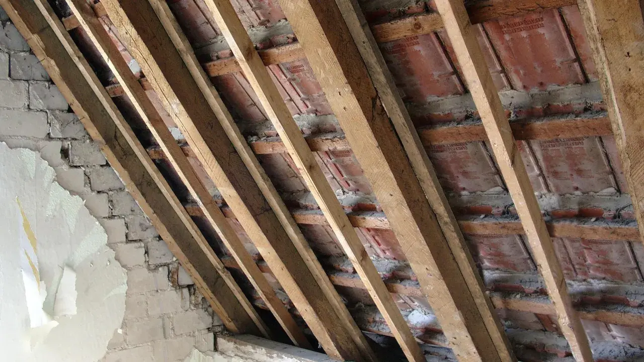Charpente en bois avec des tuiles rouges et un mur en briques décrépit. Les contre latte sont visibles, soutenant la structure.