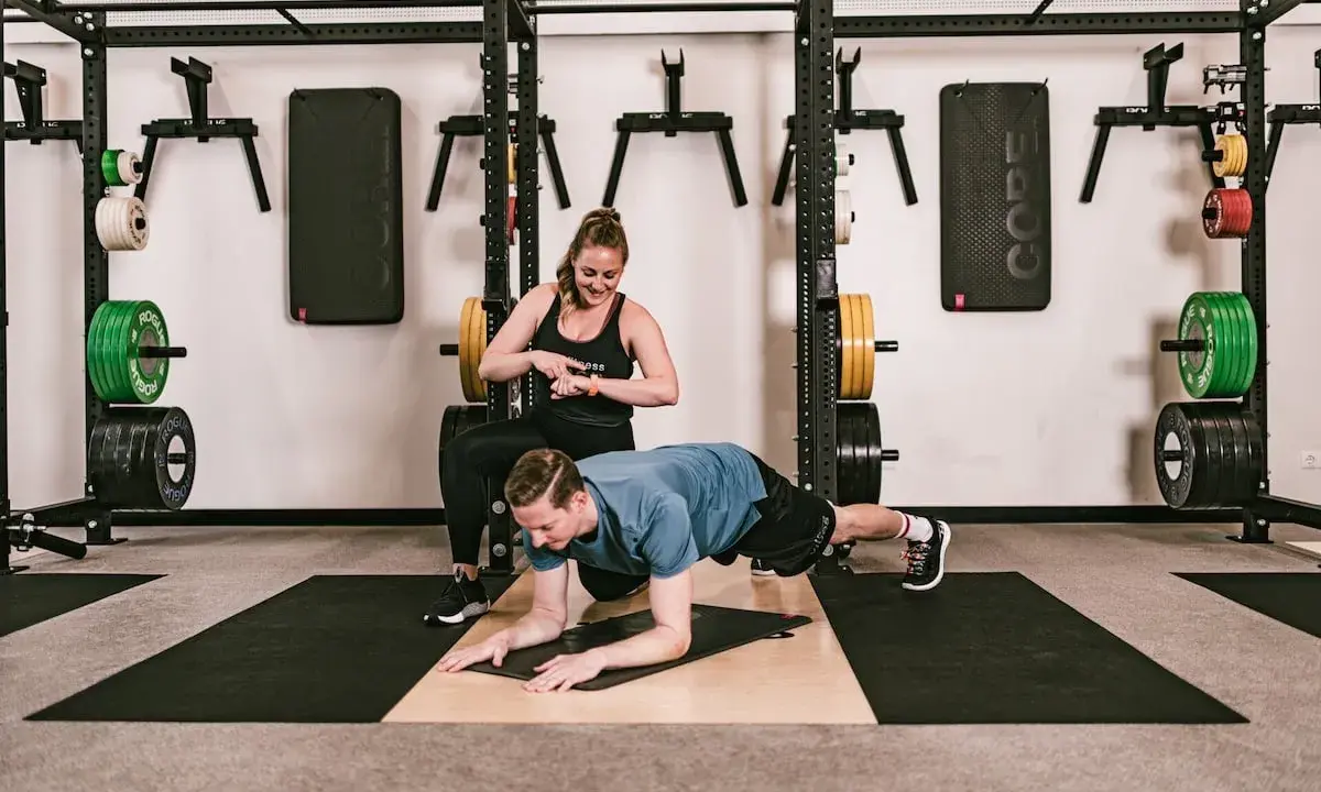 Ein Mann macht einen Unterarmstütz, während eine Trainerin die Zeit stoppt. Perfekt für einen HIIT Trainingsplan im Fitnessstudio.