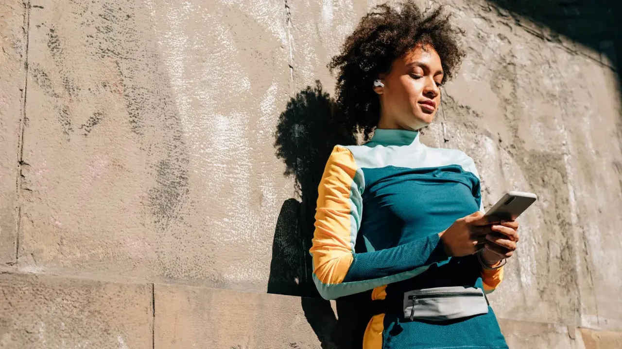 Frau mit Kopfhörern lehnt an Wand, hält handy beim joggen. Musik und Motivation für die Laufstrecke.
