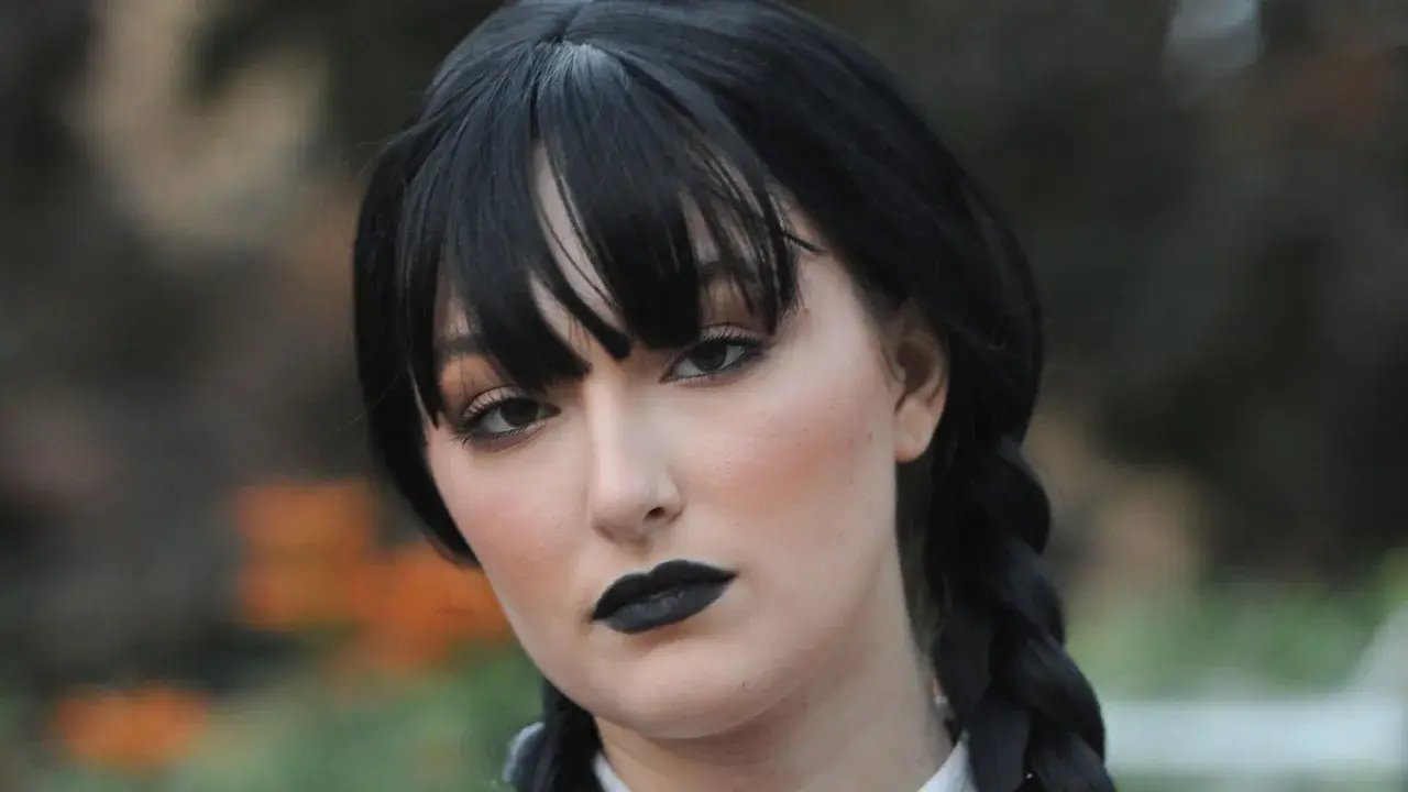Maquillaje de Miércoles Addams con labios negros y ojos ahumados, peinado con trenzas y flequillo.