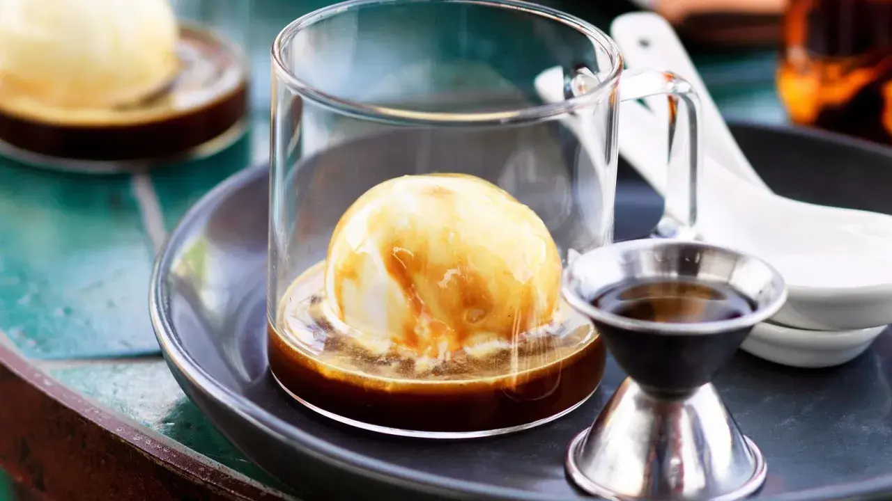 Ein cremiger Affogato mit Alkohol, serviert in einem Glas mit einer Kugel Vanilleeis, übergossen mit Espresso und einem kleinen Schnapsglas daneben.