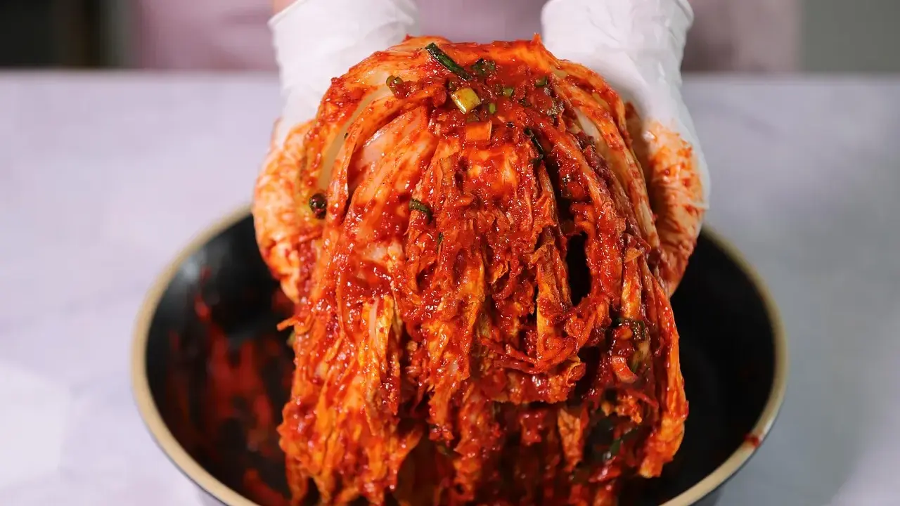 Ręce w białych rękawiczkach trzymają głowę kapusty pekińskiej pokrytą czerwoną pastą. To właśnie kimchi, tradycyjna koreańska potrawa.
