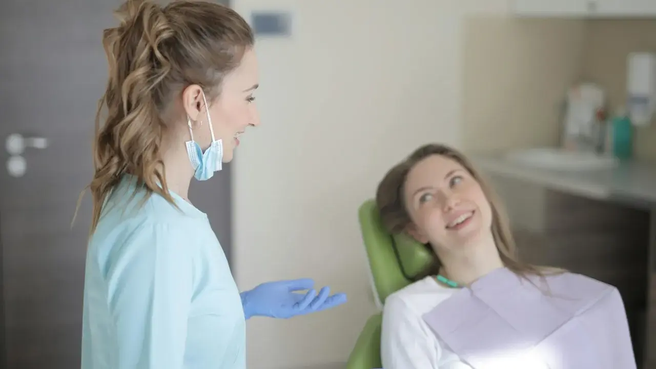 Dentystka w niebieskiej bluzie i rękawiczkach rozmawia z pacjentką. Długie studia stomatologiczne wymagają zaangażowania.