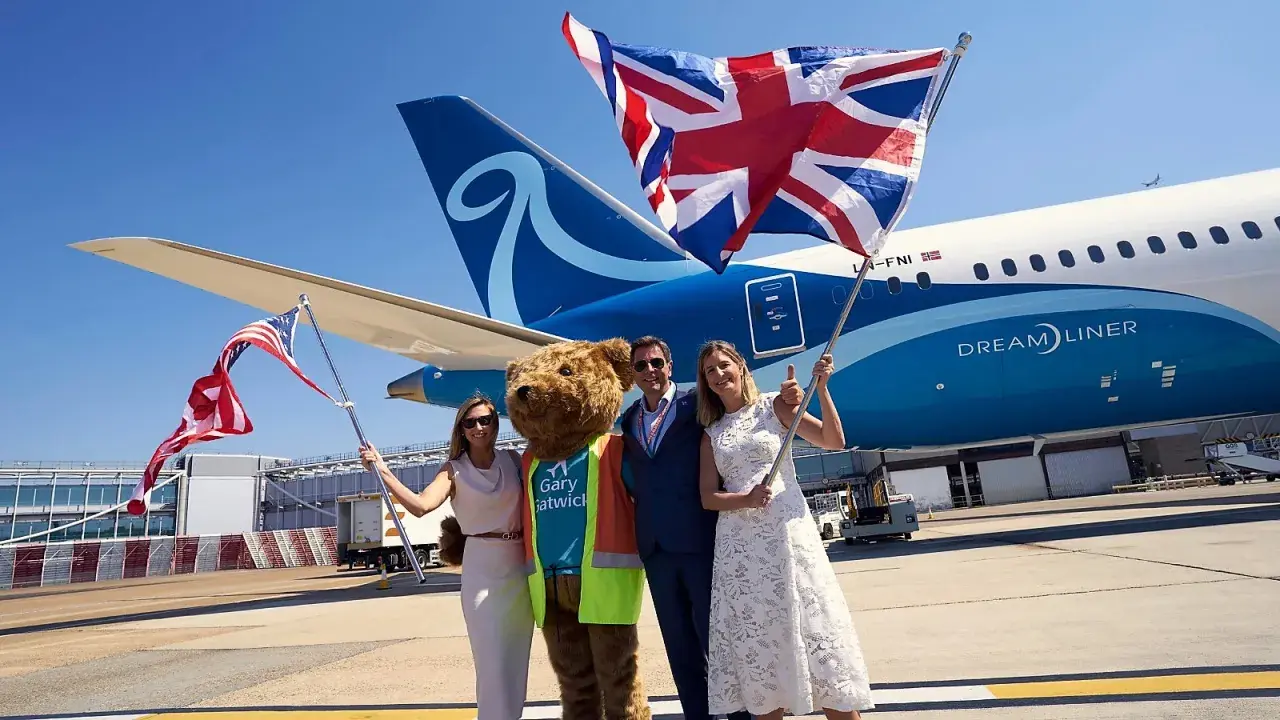 Två kvinnor och en man med en björndräkt firar framför ett flygplan från Norse Atlantic Airways. En kvinna håller en amerikansk flagga, en annan en brittisk flagga.